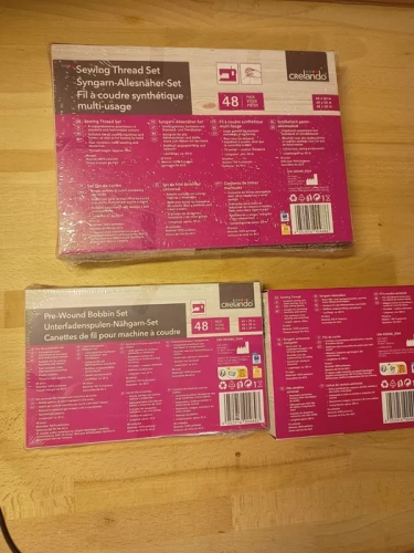 Trzy nowe oryginalnie zafoliowane zestawy nici do szycia marki Crelando w tym czterdzieści osiem szpulek czterdzieści osiem bębenków dolnych i dziesięć dużych szpul na drewnianym blacie
