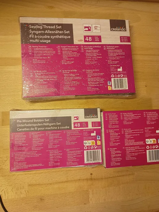 Trzy nowe oryginalnie zafoliowane zestawy nici do szycia marki Crelando w tym czterdzieści osiem szpulek czterdzieści osiem bębenków dolnych i dziesięć dużych szpul na drewnianym blacie